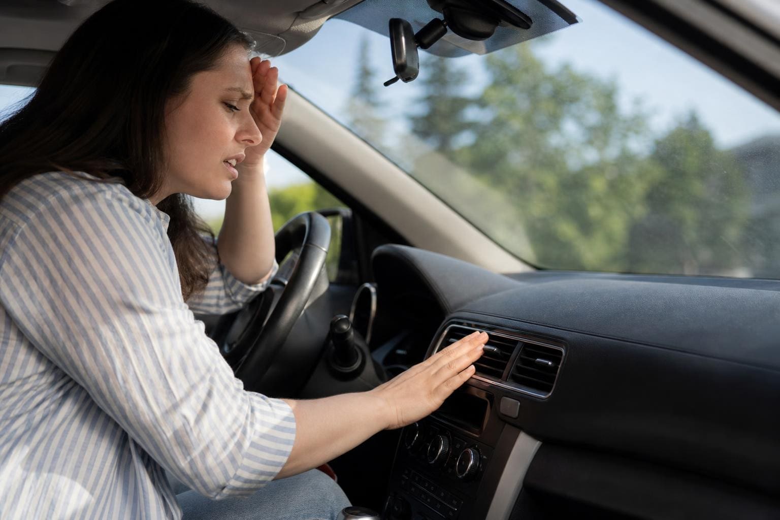 Frustrated woman sitting in a car, touching her forehead and reaching toward the dashboard air vent to check for cool air.